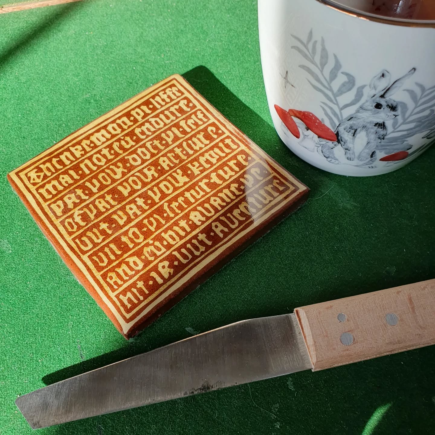 I'm prepping pages for my November class but that makes for boring photos, so enjoy this one of my desk coaster (finally removed from storage) now that I have a fancy desk to put it on. The knife is resting between page batches, as am I. 

[Image description: a medieval tile coaster in terracotta with pale yellow text on it, resting on a green baize desktop next to a mug and a cobblers knife.]