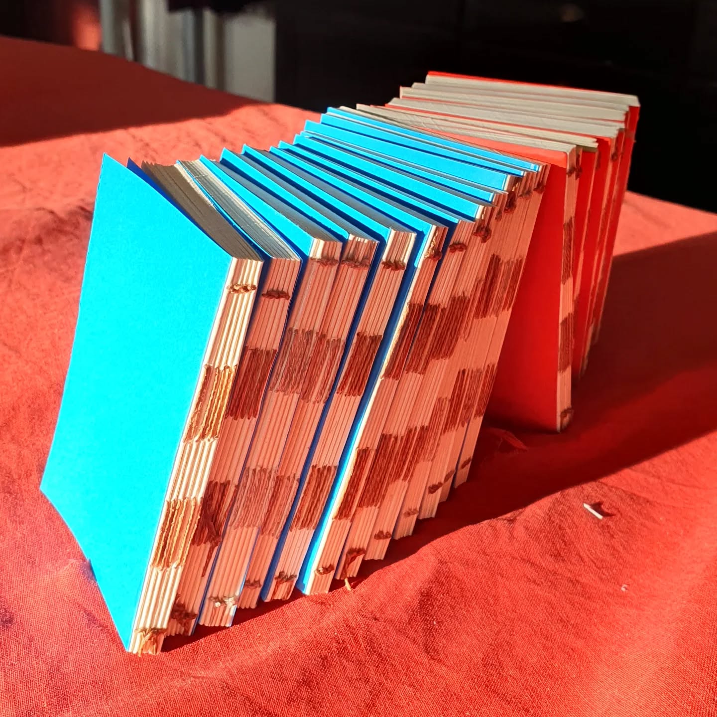 When the golden hour hits and even the dullest of shots looks fab. Teeny pocket notebooks in progress awaiting their covers.

[Image description: a stack of books some with bright teal and some with red endpapers on a bright red cloth, shining in the sunlight.]
