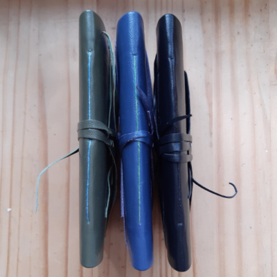The spine of three notebooks turned to show the thread colour, green book with blue thread, blue book with green thread, black book with black thread.