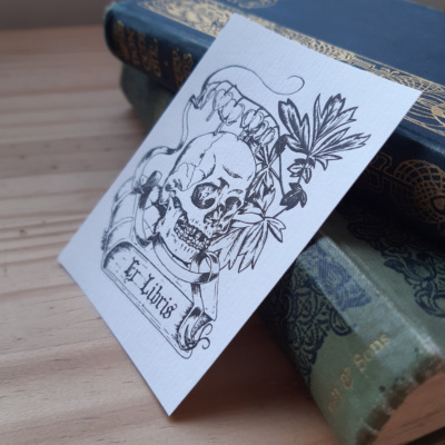 A sticker with a skull lying on a book surrounded by bleeding heart flowers. The book reads Ex Libris.