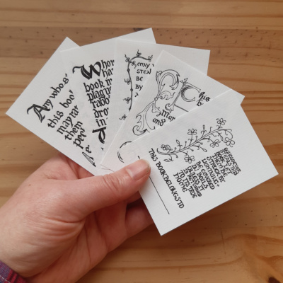 A hand holding five bookplates of different designs, each with a book curse and medieval style marginalia on.