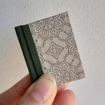 a miniature book with a green bookcloth spine and a beige paper cover with mandala like linework on.