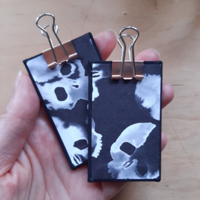 A pair of miniature clipboards held in a hand with black bookcloth on the reverse and stylised skull design on the front. Both have rose gold clips.