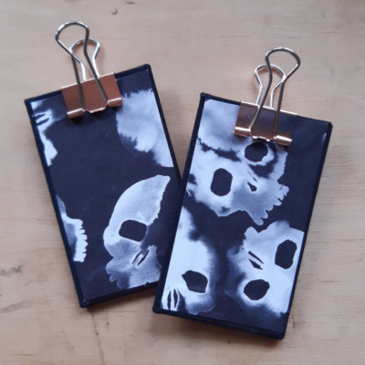A pair of miniature clipboards with black bookcloth on the reverse and stylised skull design on the front. Both have rose gold clips.