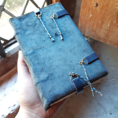A hand holding a blue grey velvet book up to the light in a window. The book has four leather straps with brass loop ends.
