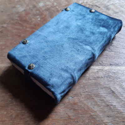 a bluegrey velvet book showing the back of the cover where the leather straps attach with four brass tacks, and the visible ridges of the cords on the spine.