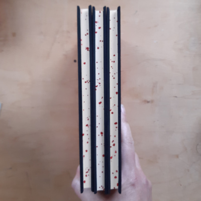 Three books being held to show red sprinkled edges, reminiscent of blood spatter