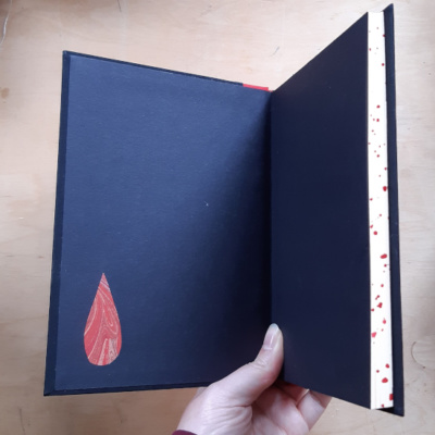 A notebook held open to show black endpapers with a blood drop insert of marbled paper.