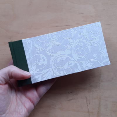 A hand holding a landscape format book with white shimmery foliage textureed paper with green bookcloth spine.