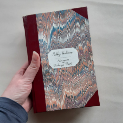 A hand holding a book with leather spine and corners, marbled paper covers and a label that reads: Mudfog Workhouse, Admissions and Discharge Book.