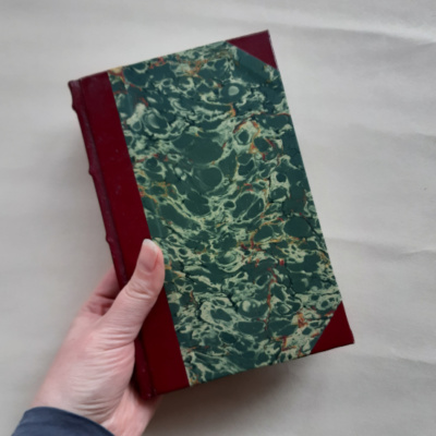A hand holding a book with maroon leather spine and corners and a green and gold marbled paper cover.