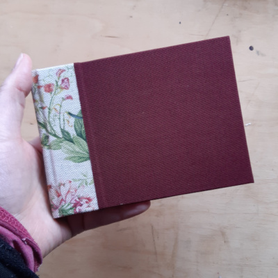 A wine red book with watercolour style floral fabric spine., held in one hand.