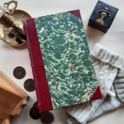 A book with maroon spine and green marbled covers surrounded by a candlestick, ink, fingerless gloves, Victorian pennies, a writing desk and a sprig of dried pine cones.,
