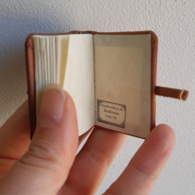 a hand holding a miniature binding open to show off the endpapers and bookbinder's ticket with the name of the bindery and place of binding on it.