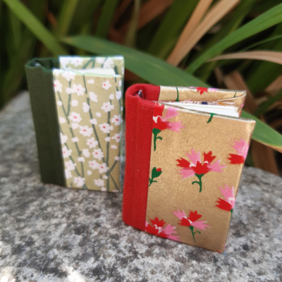 two floral themed miniature notebooks, one in shades of green with white flowers, one in red and gold with red and pink flowers.