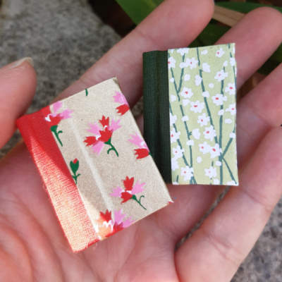 a hand holding two floral themed miniature notebooks, one in shades of green with white flowers, one in red and gold with red and pink flowers.