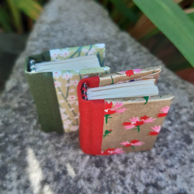 two floral themed miniature notebooks, one in shades of green with white flowers, one in red and gold with red and pink flowers. photo taken from above to show the colour matched endbands