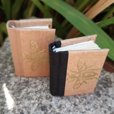two miniature books with gold bees on the covers one has a black cloth spine one a sand coloured cloth spine