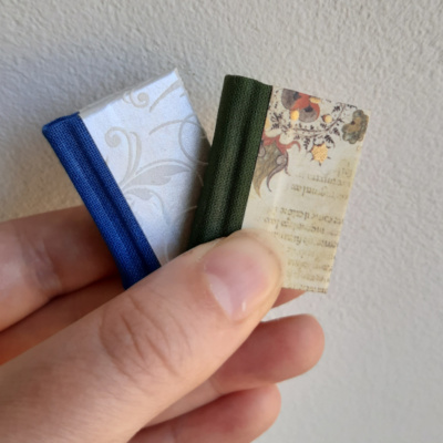 a hand holding two miniature books one with a blue spine and white swirled paper, the other with a green spine and illuminated manuscript paper designs on the cover.