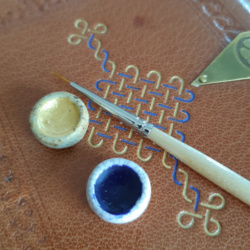 a close-up shot of a paint brush and two paint palettes resting on a decorated leather book with gold and blue painted tooling.