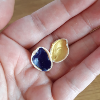 a hand holding two pistachio shells with paint in, one has blue paint, one has gold paint.