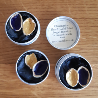 Three tins holding two pistachio shells each, one with blue paint one with gold paint, on a bed of black tissue paper. a lid next to it reads "Ultramarine Blue & Gold Paint (vegan-friendly) In pistachio shells"