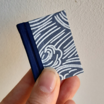 A miniature book with a blue cloth spine and blue and white wave patterned covers held by between finger and thumb