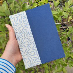 a hand holding an A5 book with a cream and blue foliage fabric on the spine and dark blue cloth covers.