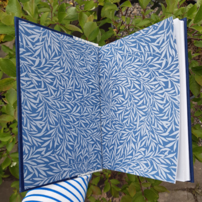 A hand holding an A5 book open to show off endpapers made of vintage paper with a willow-leaf pattern in two shades of blue.