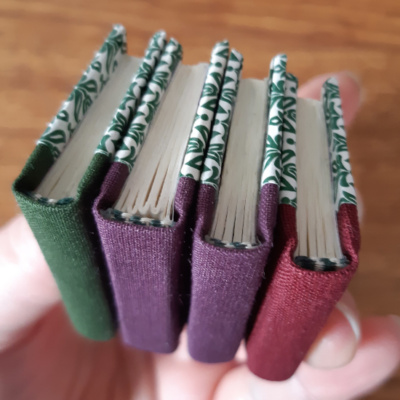 four miniature books held up to show the spines that are green, purple, purple, and wine-red.