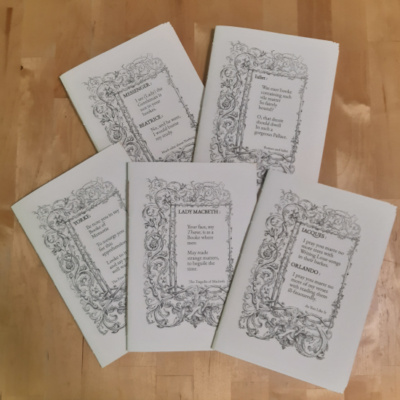 5 A5 notebooks with white covers lying on a wooden table. Each notebook has the same foliage border on it, the centre of which has a different Shakespeare quote in it.