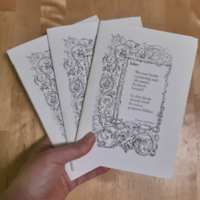 A hand holding 3 A5 notebooks with white covers. Each notebook has the same foliage border on it, the centre of which has a Shakespeare quote in it.