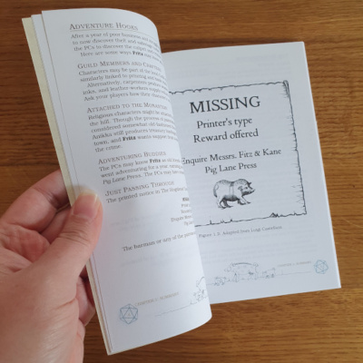 An open RPG adventure booklet on a wooden background. The image visible on the open page reads "Missing: Printer's Type, Reward Offered".