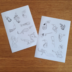 Two sticker sheets on a wooden background. The stickers are black and white and in the shape of hands or creatures pointing. They include a hand emerging from a shell, a man and woman pointing, a knight with a tail, and dragon bodied hands.
