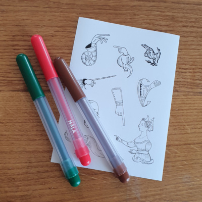 A sticker sheet on a wooden background. The stickers are black and white and in the shape of hands or creatures pointing including a lady a snail hand and a dragon hand. 3 felt tip pens in brown, green, and red are lying on top.
