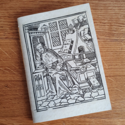 A pamphlet notebook lying on a wooden background. It has a parchment effect card cover, with a woodcut image of a bored looking man sat at a desk surrounded by books.