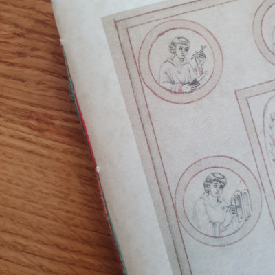 A close-up photo of a section of a notebook cover image showing two roundels of a medieval manuscript drawing. One roundel has a monk cutting a quill, the other has a monk marking up in a wax tablet.
