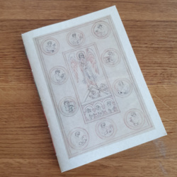 An A5 notebook lying on a wooden table, the notebook has parchment card covers and the image is of a medieval manuscript image of archangel Michael in a rectangular frame, surrounded by 10 roundels, each with a monk doing a stage of manuscript production.