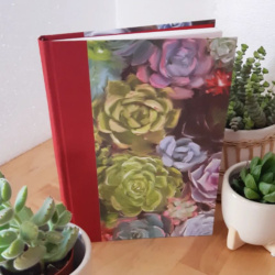 A book surrounded by succulents. It has a red book cloth spine and the cover has a watercolour style image of succulents on it.