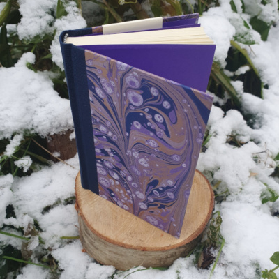A book stood open on a tree stump surrounded by snow and foliage. The book has a dark blue spine and covers covered in purple and gold swirled marbled endpapers with white speckles. The endpapers are a matched purple to the marbling.