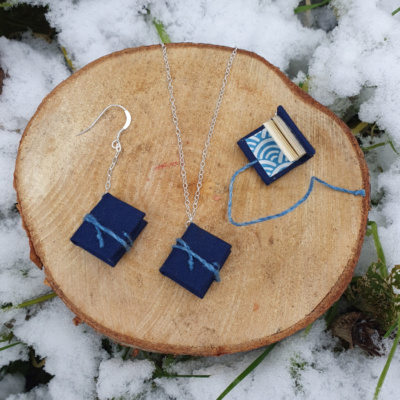 Three miniature books with blue book cloth covers, and blue ties. Two are on chain drop earrings, one is hung on a necklace. One earring book is open to show off the white and blue wave patterned endpapers.