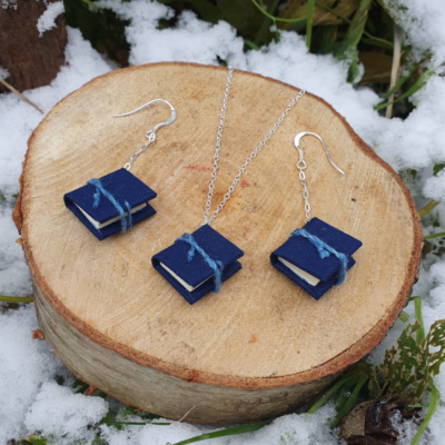 Three miniature books with blue book cloth covers, and blue ties. Two are on chain drop earrings, one is hung on a necklace. They are displayed artfully on a cut log surrounded by snow and foliage.