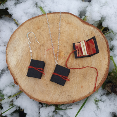 Three miniature books with black book cloth covers, and red ties. Two are on chain drop earrings, one is hung on a necklace. One earring book is open to show off the red and gold marbled endpapers.