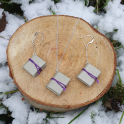 Three miniature books with shimmery silver book cloth covers, and purple ties. Two are on chain drop earrings, one is hung on a necklace. They are displayed artfully on a cut log surrounded by snow and greenery.