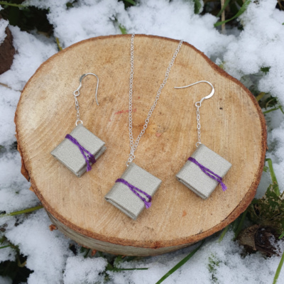 Three miniature books with shimmery silver book cloth covers, and purple ties. Two are on chain drop earrings, one is hung on a necklace. They are displayed artfully on a cut log surrounded by snow and greenery.