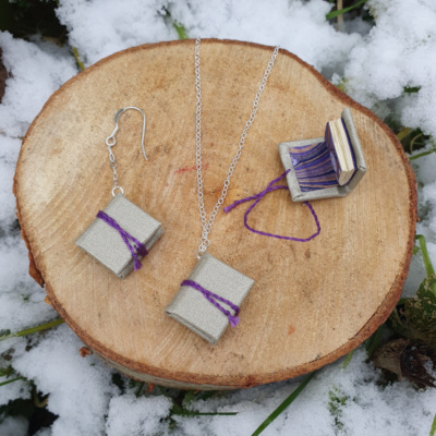 Three miniature books with shimmery silver book cloth covers, and purple ties. Two are on chain drop earrings, one is hung on a necklace. One earring book is open to show off the purple and gold marbled endpapers.