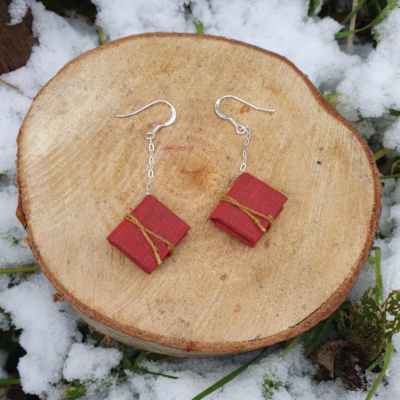 A pair of red miniature books with golden yellow ties, are hung from silver ear hooks and chain. They are displayed artfully on a cut log surrounded by snow and greenery.