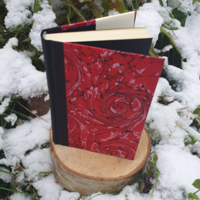 A book stood open on a tree stump surrounded by snow and foliage. The book has a black spine and covers covered in black and red swirled marbled paper. The endpapers are a matched black to the marbling.