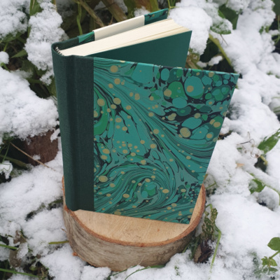 A book stood open on a tree stump surrounded by snow and foliage. The book has a dark green spine and covers covered in green swirled marbled paper with gold speckles. The endpapers are a matched green to the marbling.
