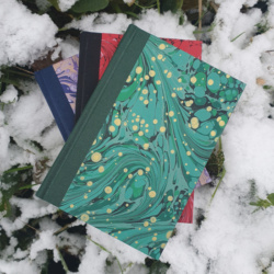 Three books with book cloth spines and marbled paper covers in different colours lying surrounded by snow and foliage